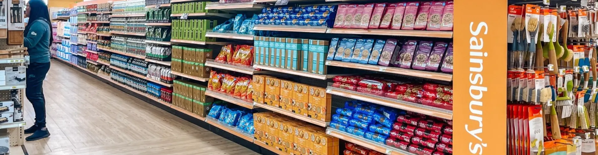 A person in a hooded jacket browses the aisles in a Sainsbury's store. Shelves display a variety of packaged snacks and groceries.