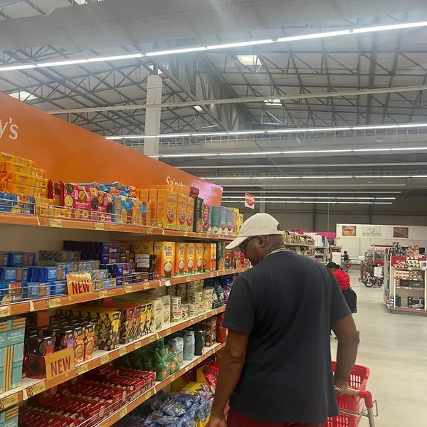 A person wearing a cap and carrying a red shopping basket is browsing the cereal aisle in a well-lit grocery store. Various cereal boxes, including brands like Kellogg's, are displayed on the shelves. A person wearing a cap and carrying a red shopping basket is browsing the cereal aisle in a well-lit grocery store. Various cereal boxes, including brands like Kellogg's, are displayed on the shelves.