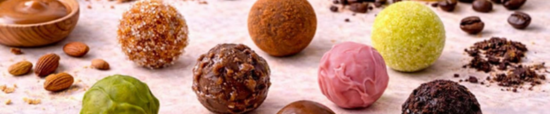 A variety of colorful truffles arranged on a surface, surrounded by almonds, coffee beans, and a wooden bowl containing a brown substance. A variety of colorful truffles arranged on a surface, surrounded by almonds, coffee beans, and a wooden bowl containing a brown substance.