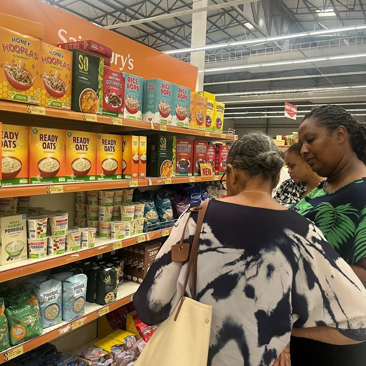 Three people stand in front of a grocery store shelf stocked with various breakfast cereals and porridge, including brands like Three people stand in front of a grocery store shelf stocked with various breakfast cereals and porridge, including brands like