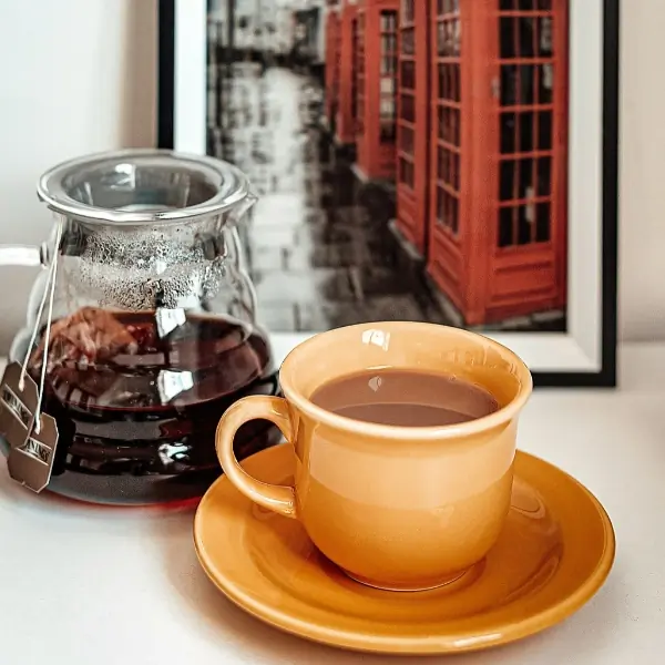 An orange tea cup stands in front of a glass tea pot and a framed picture of the red London telephone booths. Image credit: Cristiane Teston sourced on Unsplash royalty free images.